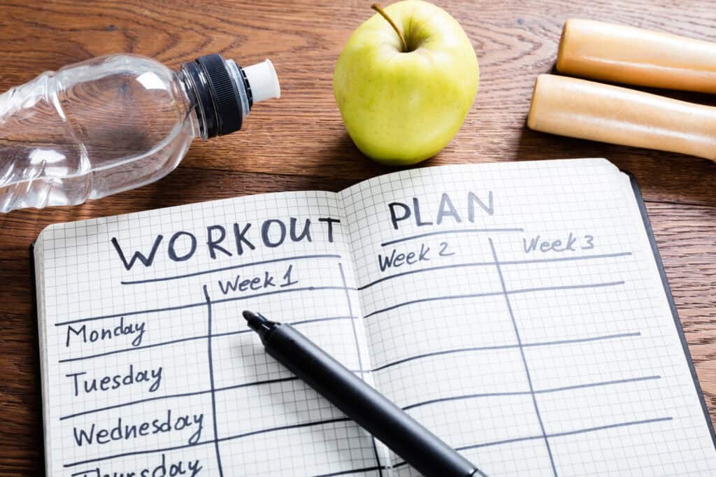 Open notebook showing a weekly workout split plan with a pen, water bottle, and apple