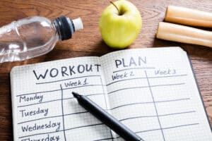Open notebook showing a weekly workout split plan with a pen, water bottle, and apple